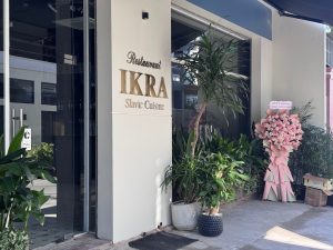 Exterior view of IKRA Slavic Cuisine restaurant with glass doors, pale wall, and potted plants outside. a large pink floral standing arrangement near the entrance