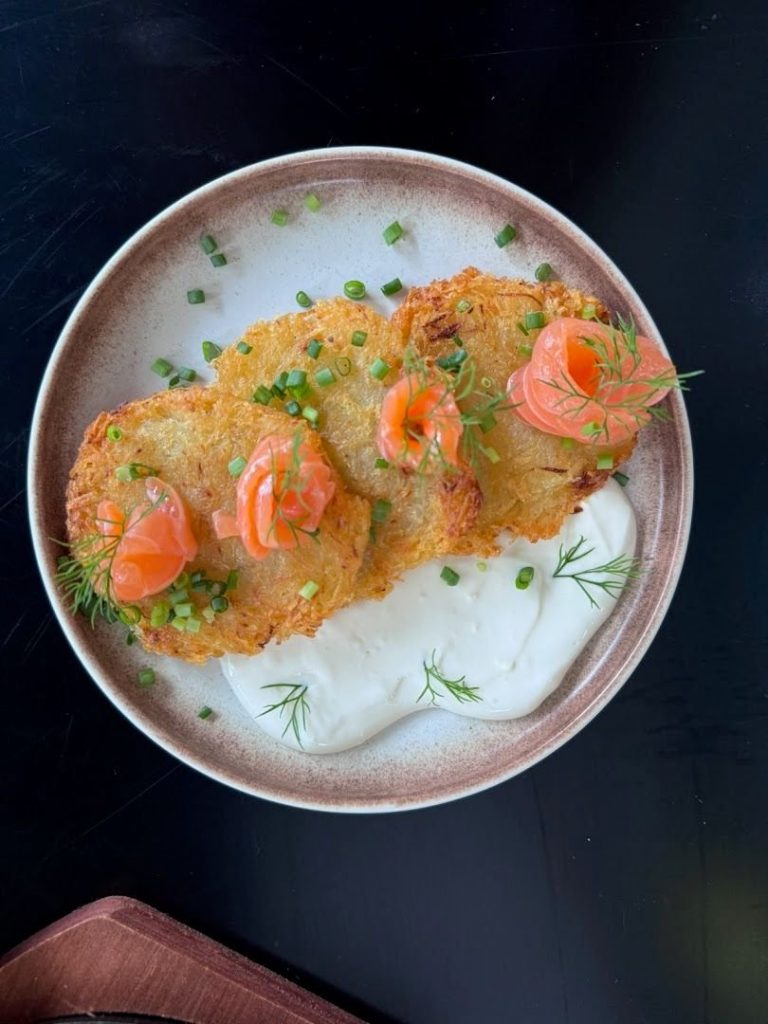 Crispy potato rosti slices topped with salmon rose garnished with chives on a beige plate with dill sprigs and a dollop of cream sauce on the side
