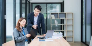 Two colleagues collaborating at a desk in a modern office, with a laptop, clipboard, and stacked papers on the table.