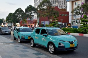 Two turquoise taxis lined up along a city street, with a modern red-brick building and trees in the background.