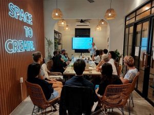 Group of people sit around a long table in a conference room, presenting to a screen as neon letters read 'SPACE TO CREATE' on the wall.