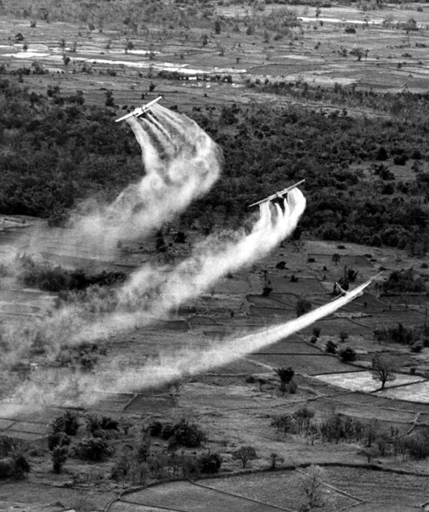 Three planes drop water in formation, releasing huge white plumes over a rural, tree-filled landscape.