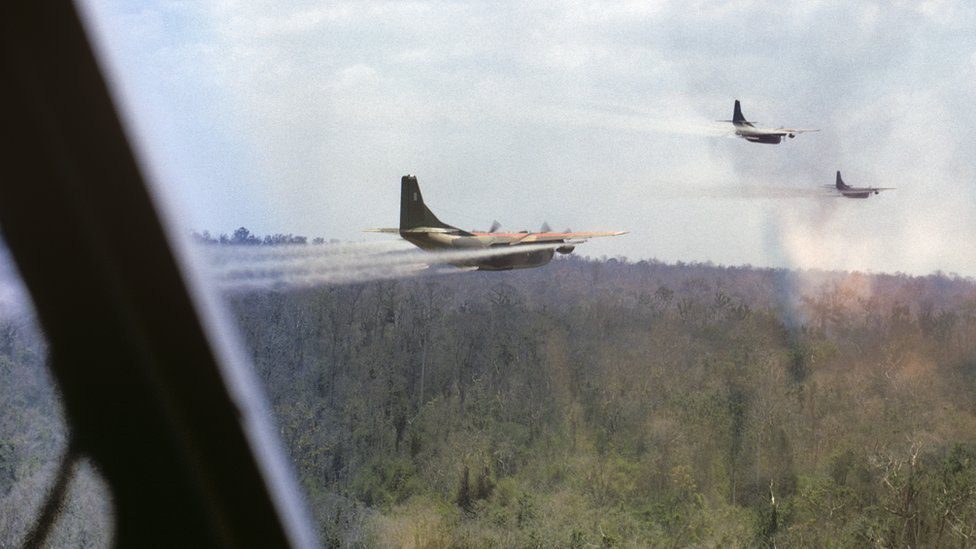 Three firefighting planes fly in formation over a forest, releasing retardant spray toward a wildfire area. (View from a cockpit)