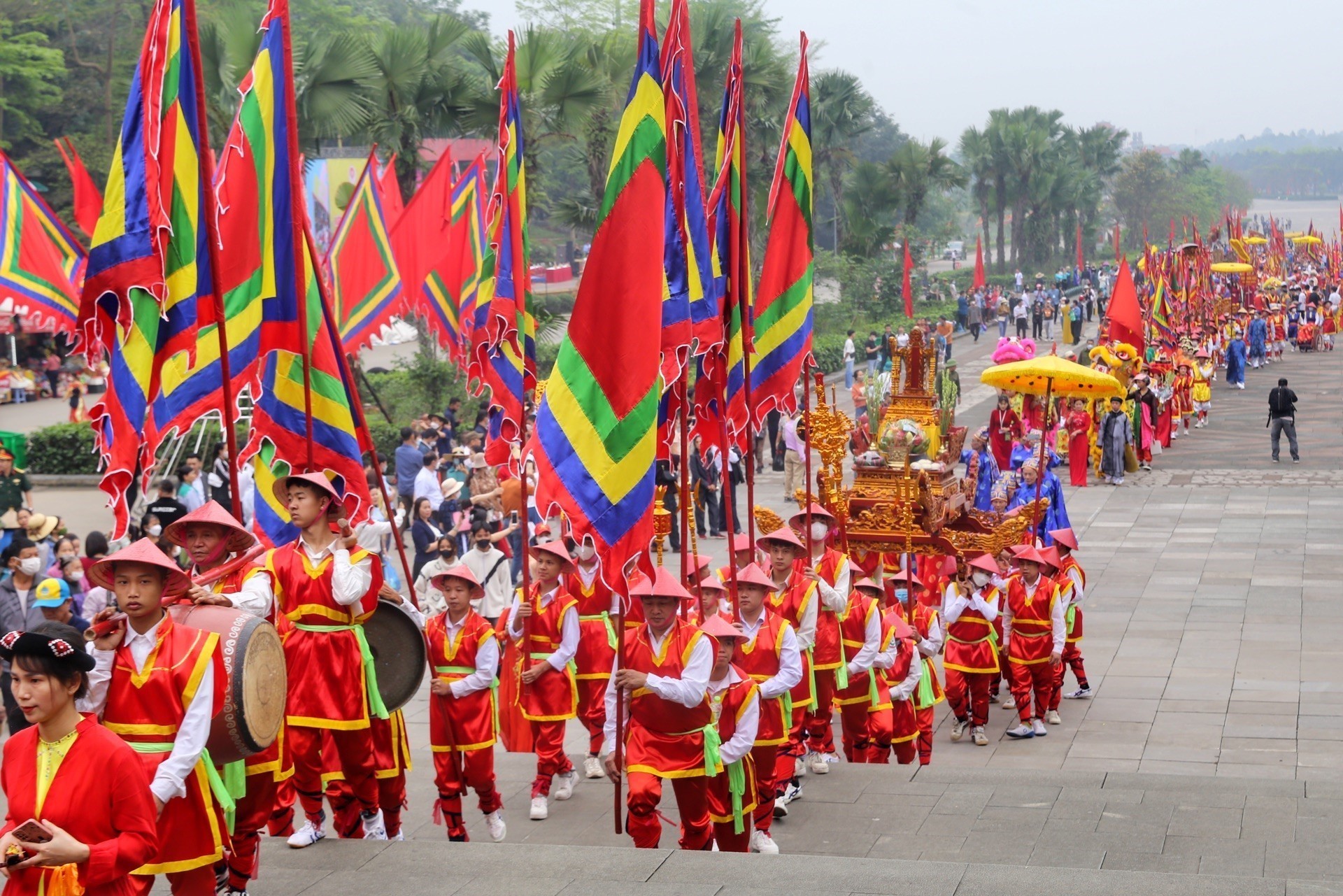 Vibrant parade with flag-bearers in red and gold costumes carrying colorful, striped flags down a wide street.
