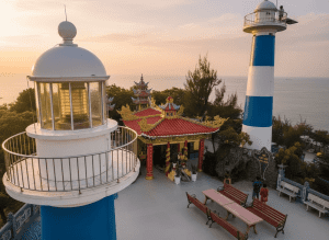 Sunset view of a seaside lookout with a white domed lantern and a blue-and-white striped lighthouse beside a colorful Asian temple with red roofs.