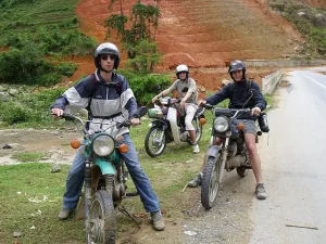 Three motorcyclists wearing helmets rest on their bikes on a rural roadside with green hills and orange soil in the background.