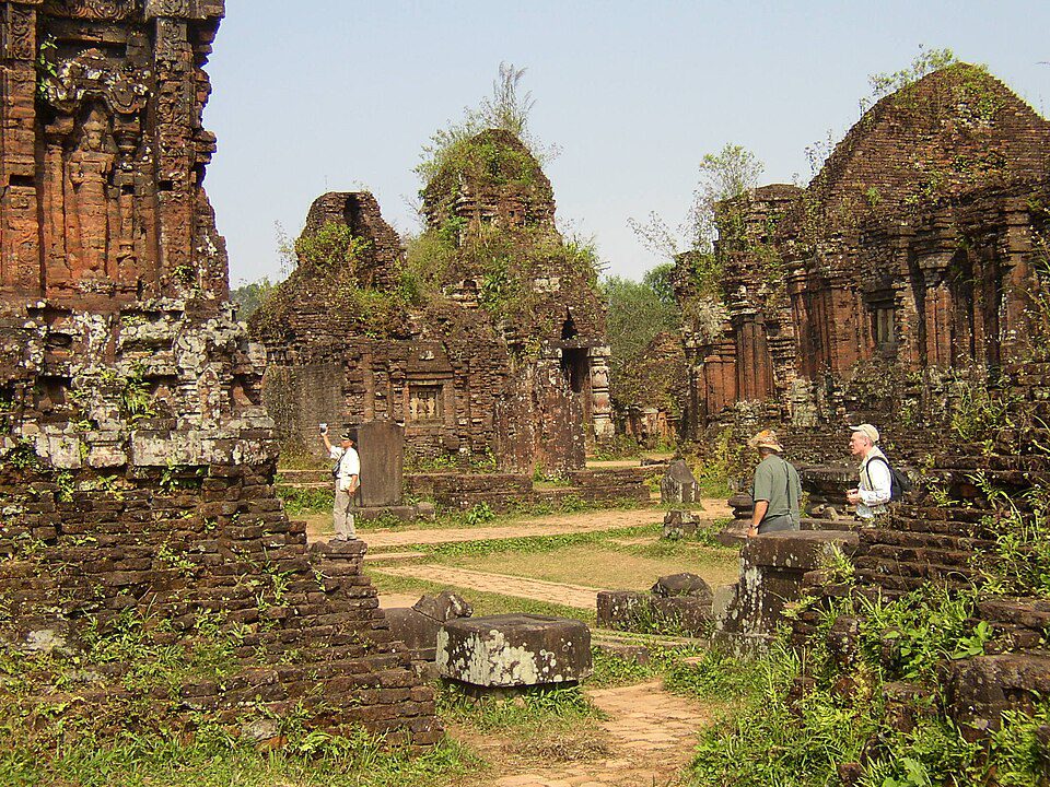 Ancient stone temple ruins with plants growing on top; three visitors walk among the structures.
