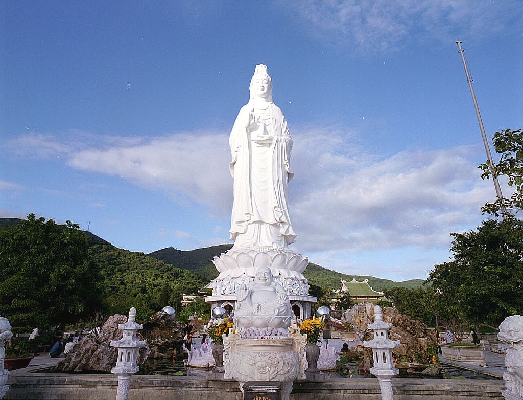 Statujë e madhe e bardhë e Bodhisattva-s në këmbë mbi një piedestal zambaku në një kopsht tempulli.