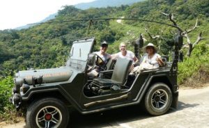 Three tourists ride in a vintage olive-green open-top jeep along a mountain road with green hills in the background, smiling at the camera.