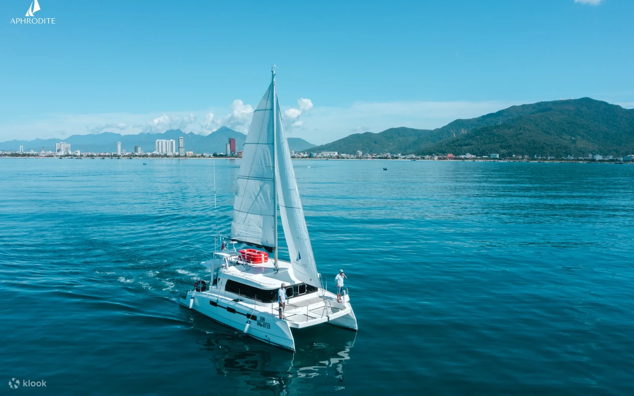 White catamaran sailing on calm blue water with a distant coastline of high-rise buildings and green mountains under a clear sky (Aphrodite branding visible in the top-left).