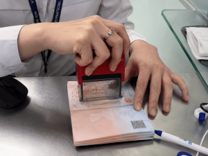Close-up of hands stamping a passport on a metal counter, with a ring on the finger and a lanyard in view.