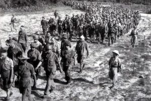 Large group of soldiers marching in a long column across a dusty field, led by a few officers.