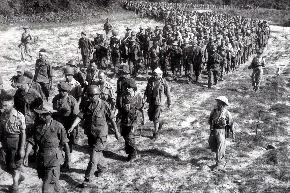 Large group of soldiers marching in a long column across a dusty field, led by a few officers.