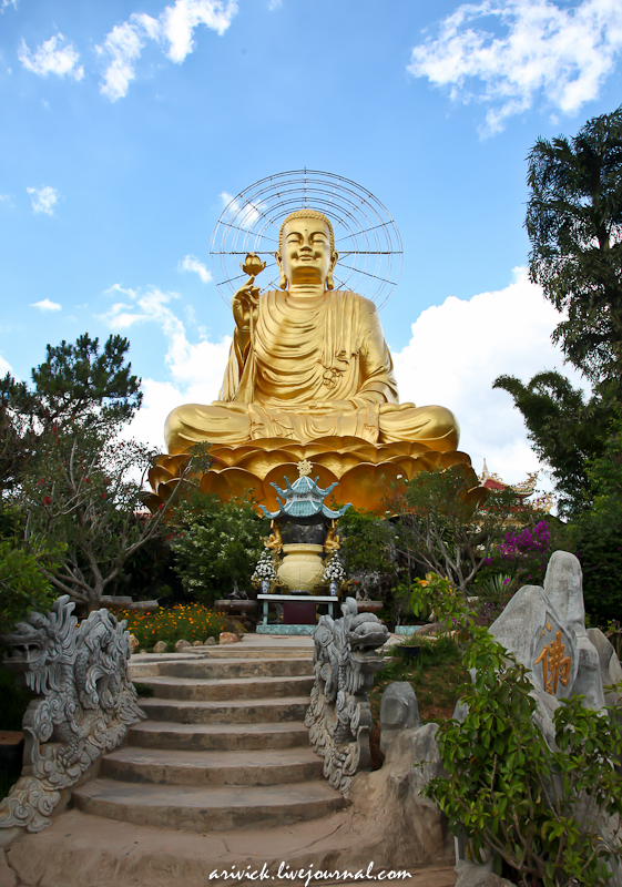 Large gold Buddha statue seated in a garden with a radiant halo, blue sky behind, stone steps and carved dragon railings leading to an incense burner shrine.