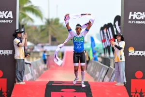 Male athlete crosses finish line with arms raised in victory, ribbon in hand, blue-and-white kit amid cheering volunteers and sponsor banners.