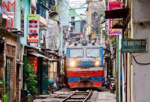 A blue and red locomotive passes down a narrow street with shops on both sides, overhead wires crisscrossing the alley.