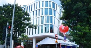 Modern curved white glass building on a tree-lined street, with a white tent set up below and a large red promotional balloon nearby.