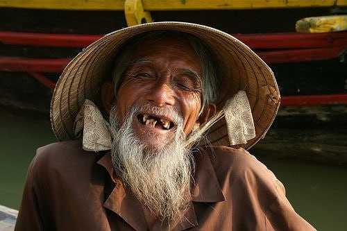 Smiling elderly man with a long white beard and missing teeth, wearing a wide-brimmed hat and brown jacket, in front of a colorful boat.