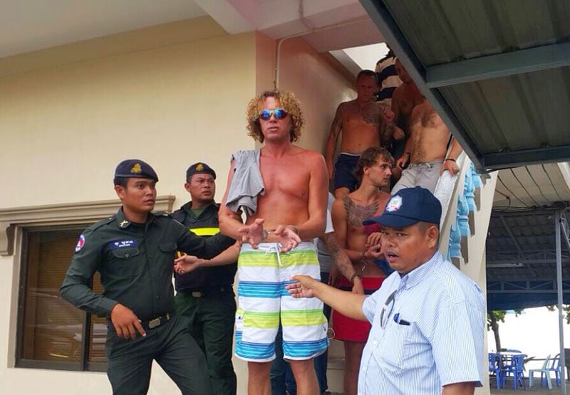 Shirtless men on stairs being guided by uniformed security guards outside a building.