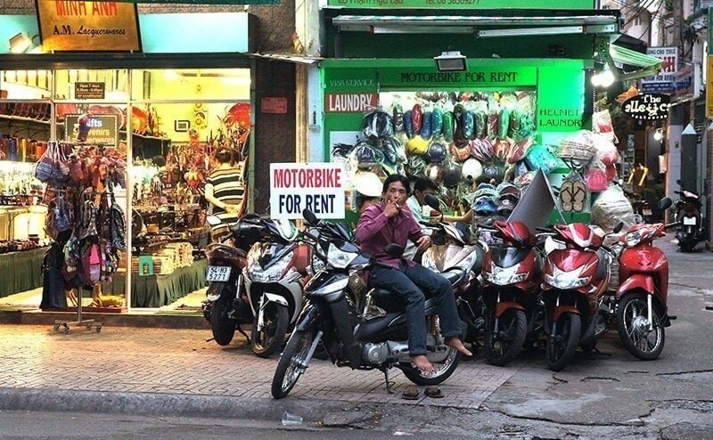Man sits on a motorbike in a row of parked motorcycles outside a shop with a 'Motorbike for Rent' sign
