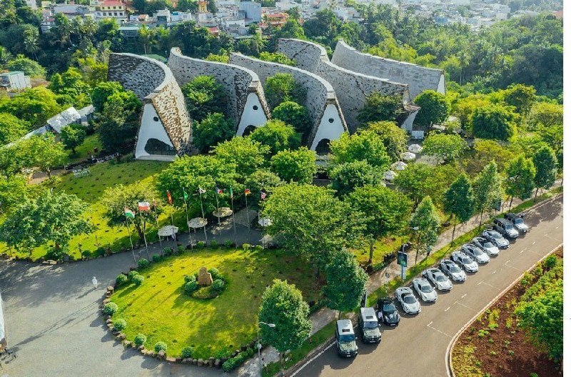 Aerial view of a park with distinctive curved stone buildings surrounded by trees, a circular grassy area, and a line of parked cars along the street