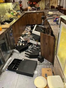 Restaurant bar area with broken appliances and scattered debris across the floor, including a tipped-over microwave and open cabinet doors amid wooden counter cabinets.