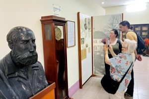 Museum visitors study display panels beside a large bust and a tall clock.