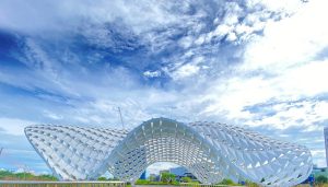 Futuristic white lattice arch building with wing-like curved sections under a bright blue sky.
