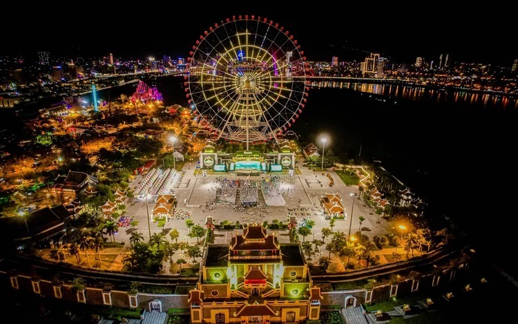 Aerial night view of a waterfront amusement park with a giant illuminated Ferris wheel and colorful lights along the shore.