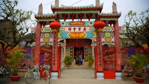 Ornate Chinese temple gate with red pillars, colorful murals, and red lanterns, bicycles parked outside amid plants and trees.