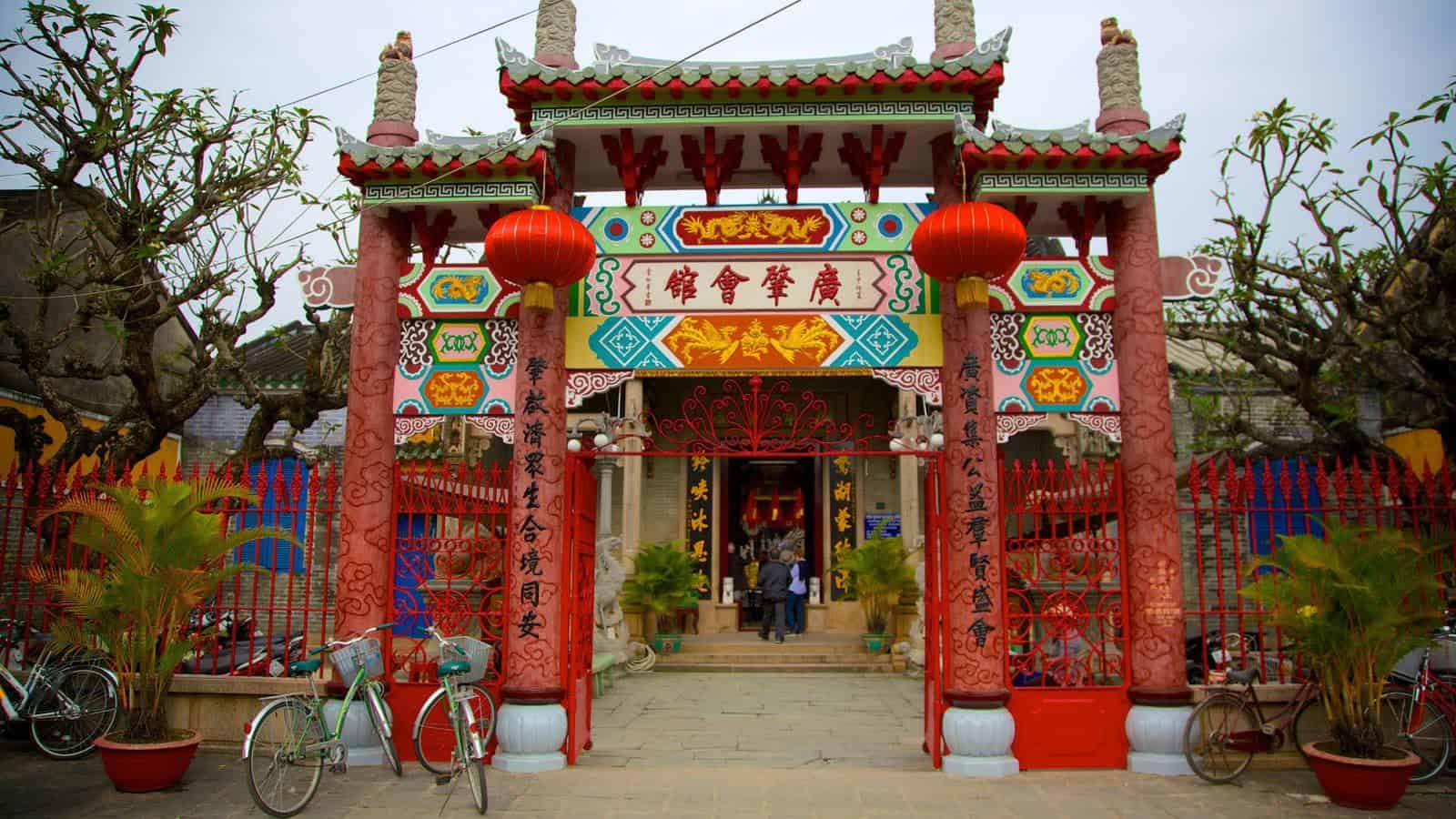 Ornate Chinese temple gate with red pillars, colorful murals, and red lanterns, bicycles parked outside amid plants and trees.