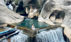 Man relaxing in a turquoise pool on a stone ledge, with a waterfall and gray canyon rocks surrounding.