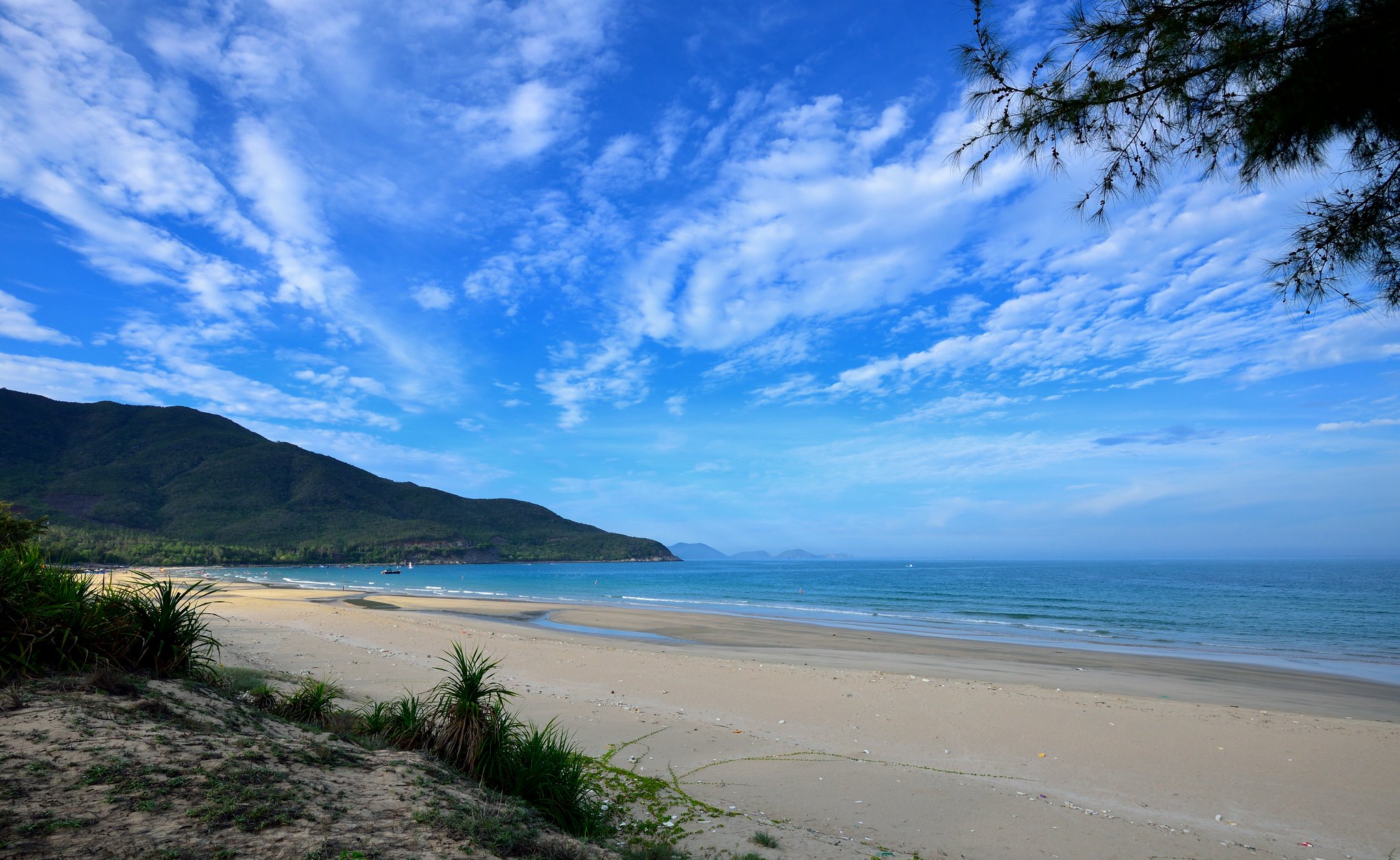 Sunny beach with calm waves, sandy shore, and green hills in the distance under a blue sky with wispy clouds.