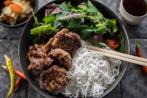 Bowl of rice vermicelli with crispy fried chicken, leafy greens, and herbs; chopsticks rest over the noodles.