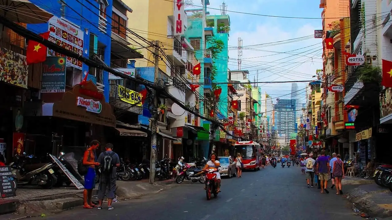 Colorful street in Vietnam with numerous flags, scooters, pedestrians, and a red bus in the distance.]** Note: remove this line
