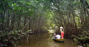 Three people ride a small yellow boat along a narrow muddy canal through dense mangrove trees, one person standing with a conical hat steering.