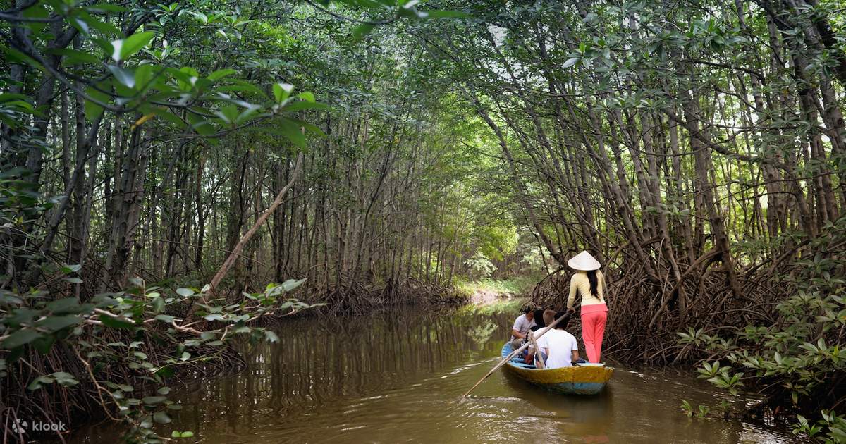 Three people ride a small yellow boat along a narrow muddy canal through dense mangrove trees, one person standing with a conical hat steering.
