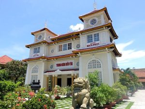 Three-story cream building with red-tiled roofs, lattice windows, potted plants, and a stone guardian lion.