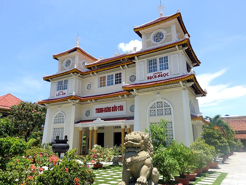 Three-story cream building with red-tiled roofs, lattice windows, potted plants, and a stone guardian lion.