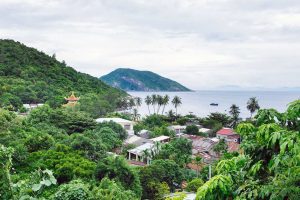 Lush tropical hillside town by the sea with palm trees, a yellow pagoda, and a calm bay with boats in the distance.