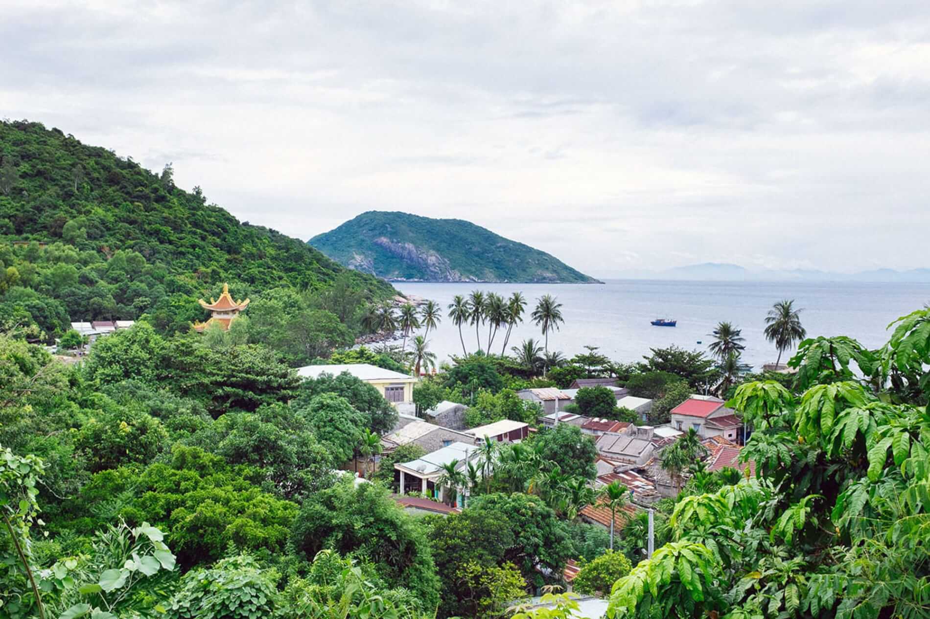 Lush tropical hillside town by the sea with palm trees, a yellow pagoda, and a calm bay with boats in the distance.
