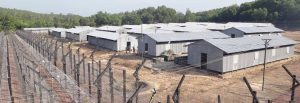 Rows of gray metal huts inside a fenced camp on dry ground, barbed-wire fencing along the perimeter with trees in the background.