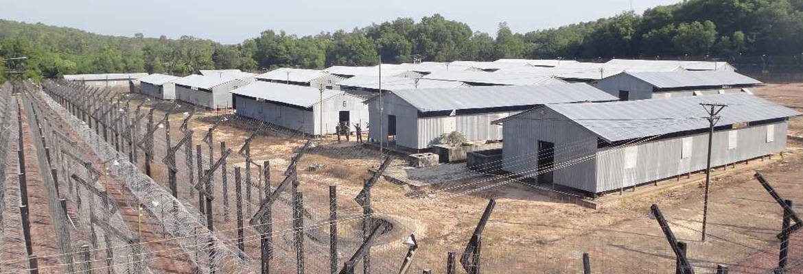 Rows of gray metal huts inside a fenced camp on dry ground, barbed-wire fencing along the perimeter with trees in the background.