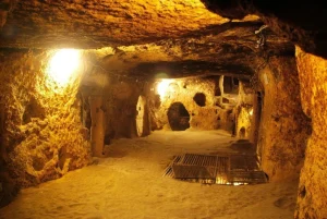 Underground cave tunnel with warm amber lighting, rough stone walls, and metal grates on the floor thinly spread over sand.