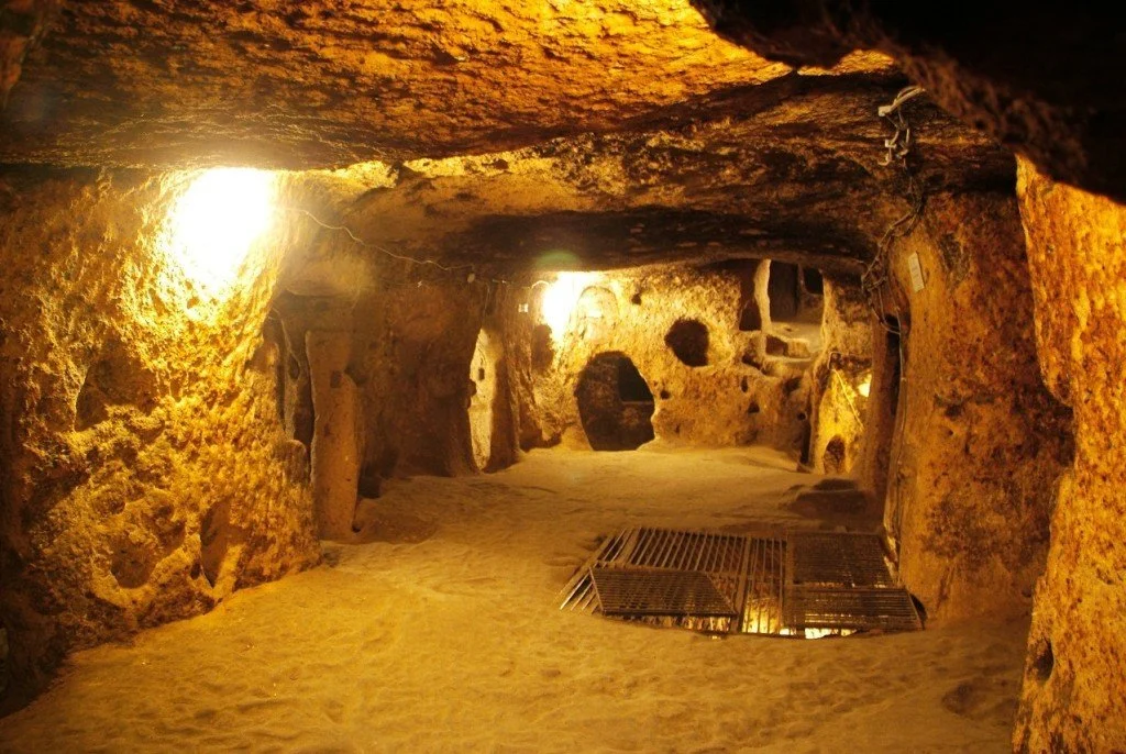 Underground cave tunnel with warm amber lighting, rough stone walls, and metal grates on the floor thinly spread over sand.