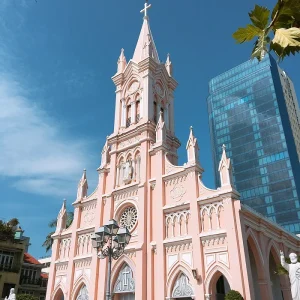 Pink Gothic church with tall central steeple and decorative arches beside a glass office building on a sunny day.