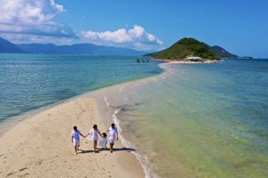 Family of four in white clothing walks hand-in-hand along a sandy beach with turquoise water and a distant green island.