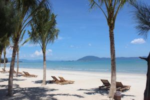 Palm trees frame a white-sand beach with wooden lounge chairs facing the calm turquoise sea and distant islands on the horizon.