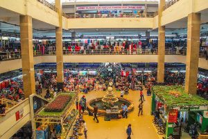 A crowded multi-level indoor market with stalls surrounding a central fountain and shoppers walking between levels.