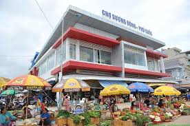 Busy outdoor market in front of a modern two-story building with red accents; stalls with colorful umbrellas and people shopping for produce.